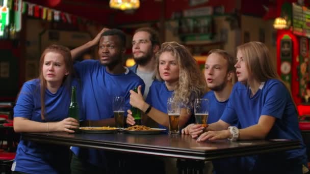 Група друзів шанувальників у блакитних футболках схвильовано дивиться матч і радіє забитому голу, стрибає і кричить емоційно. Святкувати і відзначати гол, забитий жуванням з пивом — стокове відео