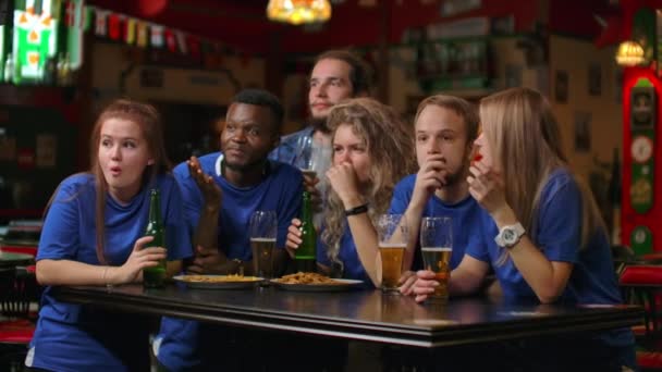 Мультиэтническая группа друзей болельщиков в синих футболках будет смотреть матч по телевизору в баре, наблюдать напряженный волнующий момент и праздновать гол. Обниматься, веселиться и делать бокал пива — стоковое видео