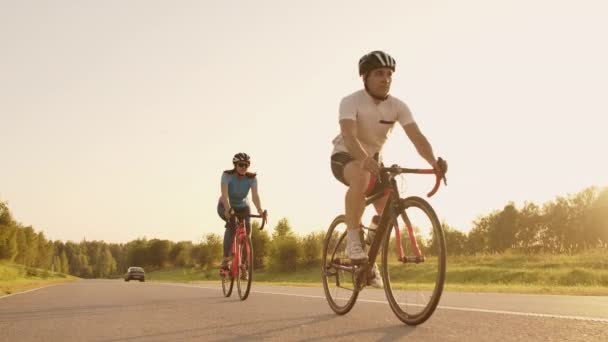 Un hombre y una mujer viajan en bicicleta deportiva en la carretera al atardecer en equipo y cascos de protección en cámara lenta 120 fps — Vídeo de stock