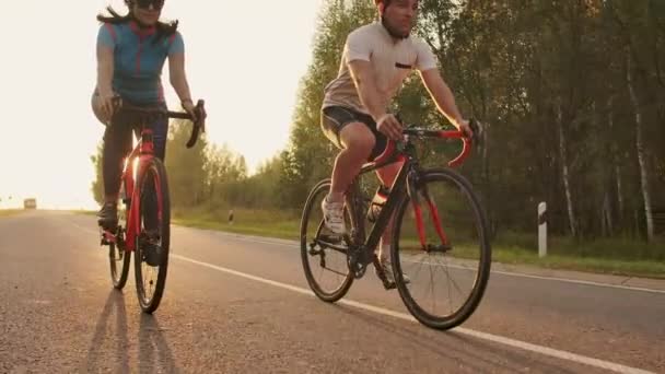 Egy férfi és egy nő a kerékpárok lovagolni az úton naplementekor együtt lassítva. A pár utazik kerékpárral. Sport kerékpáros sisakok — Stock videók