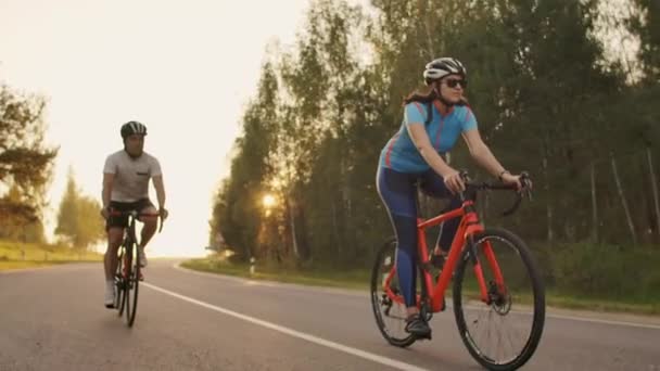 Deux cyclistes un homme et une femme roulent sur l'autoroute sur des vélos de route portant des casques et des vêtements de sport au coucher du soleil au ralenti . — Video