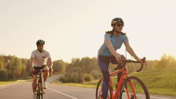 Un homme et une femme à vélo descendent la route au coucher du soleil ensemble au ralenti. Le couple voyage à vélo. Sports Casques de cyclisme — Video