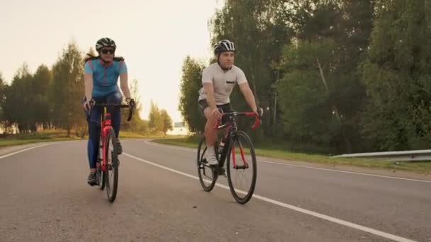 Dos ciclistas un hombre y una mujer viajan en la carretera en bicicletas de carretera con cascos y ropa deportiva al atardecer en cámara lenta . — Vídeos de Stock