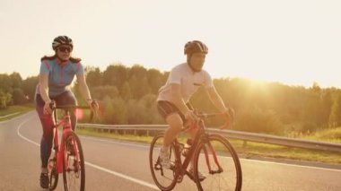 Bir adam ve bir kadın yavaş hareket 120 fps vites ve koruyucu kask gün batımında karayolu üzerinde spor bisiklet binmek