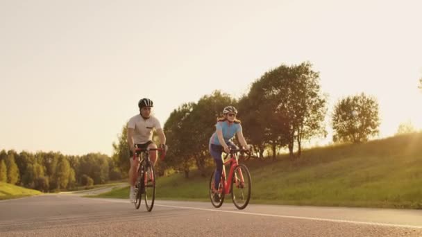 Egy férfi és egy nő a kerékpárok lovagolni az úton naplementekor együtt lassítva. A pár utazik kerékpárral. Sport kerékpáros sisakok — Stock videók