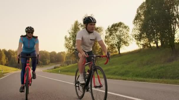 Un homme et une femme à vélo descendent la route au coucher du soleil ensemble au ralenti. Le couple voyage à vélo. Sports Casques de cyclisme — Video