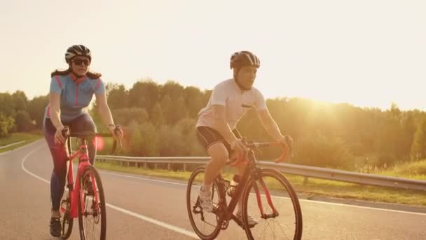 Egy férfi és egy nő lovagolni sport kerékpárok az autópályán napnyugtakor a sebességváltó és védősisakok lassítva 120 fps — Stock videók
