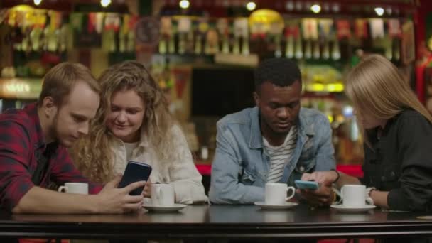 Retrato de jóvenes amigos alegres mirando el teléfono inteligente mientras están sentados en la cafetería. Gente de raza mixta sentada en una mesa en un restaurante usando teléfono móvil . — Vídeos de Stock