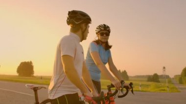Bisikletli kasklı bir adam ve bir kadın gün batımında durup konuşurlar. Otobanda bisiklete bindikten sonra dinlenin. Bisikletleri takip et. Çift sporları.