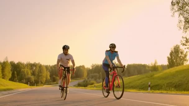 Двое велосипедистов мужчина и женщина едут по шоссе на дорожных велосипедах в шлемах и спортивной одежде на закате в замедленной съемке . — стоковое видео