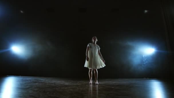 Una chica en un vestido blanco baila un ballet moderno haciendo una rotación en el escenario con humo en los focos . — Vídeo de stock