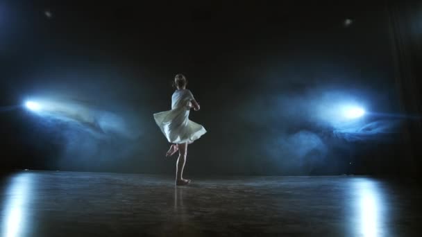 Młoda dziewczyna w białej sukni tańczy nowoczesny balet na scenie z dymem przed niebieskimi reflektorami. — Wideo stockowe