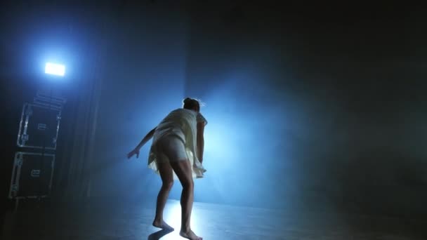 Une jeune fille en robe blanche danse un ballet moderne sur scène avec de la fumée contre les projecteurs bleus . — Video