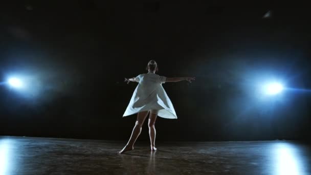 Una chica en un vestido blanco baila contemporáneo haciendo rotaciones en el escenario con humo en los focos . — Vídeo de stock