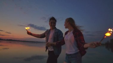 Genç aşıklar erkek ve kadın neşeyle yavaş hareket ellerinde sıcak köpüklü çürüme ile plaj boyunca çalıştırın. Romantik bir ilişki. Kıvılcımlar aşıkların ellerinde yanıyor.