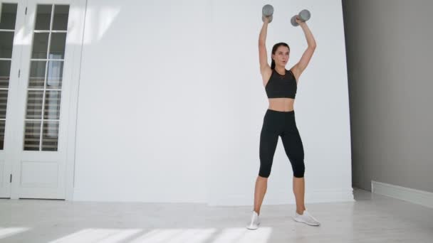 Une jeune fille brune en tenue de sport noire exécute des squats avec des haltères dans un appartement blanc par une journée ensoleillée. Mouvement lent . — Video