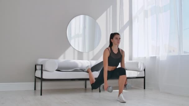 Brunette in zwarte Sportswear in een wit appartement maakt een split Squat met dumbbells in haar handen leunend op de Bank met haar voet — Stockvideo