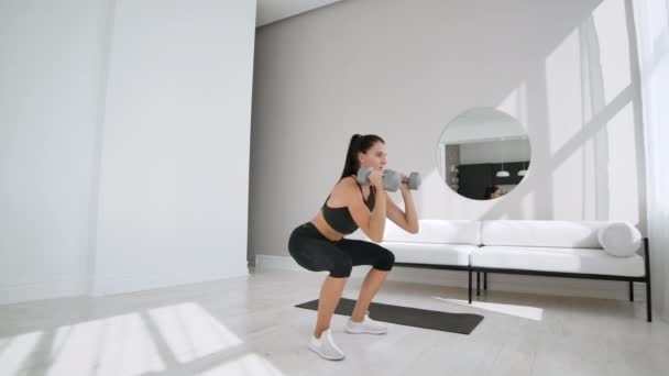 Una joven morena en ropa deportiva negra realiza sentadillas con pesas en un apartamento blanco en un día soleado. Movimiento lento . — Vídeo de stock
