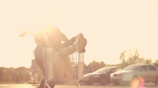 Vista lateral de una joven hembra y macho divirtiéndose al aire libre en carritos de compras. Jóvenes multiétnicos corriendo en carritos de compras. En la zona de aparcamiento con su — Vídeos de Stock