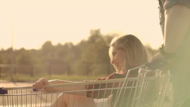 Вид сзади. Веселая молодая пара влюбленных мужчин и женщин смеются и веселятся, катаясь на телегах на парковке супермаркета в замедленной съемке на закате — стоковое видео