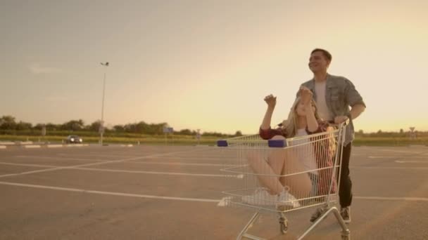 Seitenansicht einer jungen Frau und eines jungen Mannes, die sich im Freien auf Einkaufswagen amüsieren. multiethnische junge Leute, die auf Einkaufswagen rasen. auf dem Parkplatz mit ihren — Stockvideo