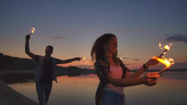 Unga älskare man och kvinna glatt springa längs stranden med varm gnistrande röta i sina händer i slow motion. Romantisk relation. Sparklers brinner i händerna på älskare. — Stockvideo