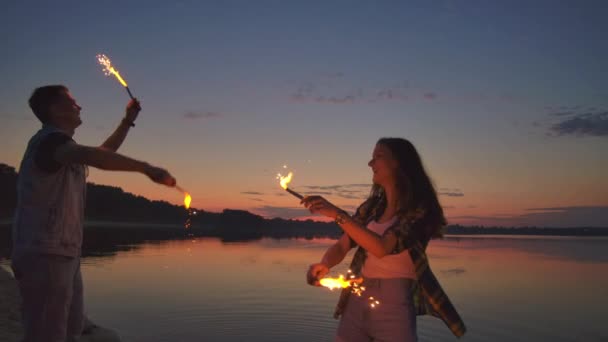 Joven pareja multiétnica feliz sosteniendo velas brillantes ardientes y corriendo por el mar durante la puesta del sol. Disparo en cámara lenta . — Vídeos de Stock