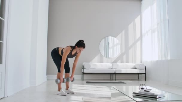 The beautiful female performs leans forward with dumbbells in her hands in the white interior of the living room. In slow motion to do exercises on the muscles of the thighs. — Stock Video