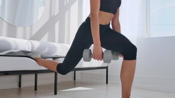 Brunetka w czarnej odzieży sportowej w białym mieszkaniu sprawia, że Split squat z hantlami w jej rękach opierając się na kanapie z nogą — Wideo stockowe