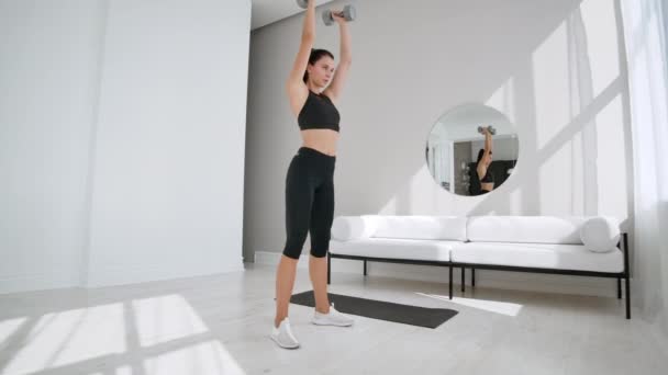 Sterk en fit mooi meisje in een atletische top doet squat oefeningen in haar lichte en ruime woonkamer met minimalistisch interieur. — Stockvideo