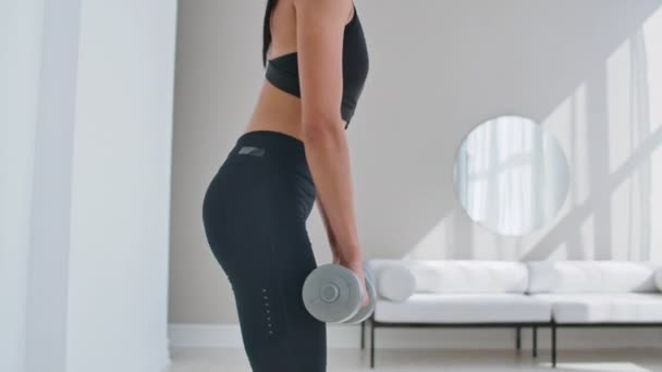 Uma jovem atlética em um apartamento brilhante realiza um deadlift com halteres em casa, fazendo inclinações para frente para fortalecer os músculos das coxas . — Vídeo de Stock