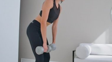 Parlak bir dairede genç bir atletik kadın evde dumbbells ile bir deadlift gerçekleştirir, ileri uyluk kasları güçlendirmek için eğilir yapma.