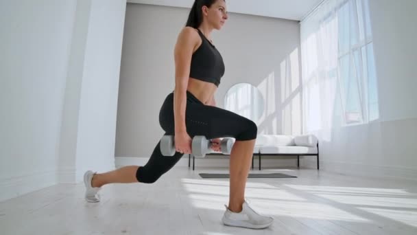 A woman at home on a Sunny day in the living room lunges back with dumbbells in her hands. Healthy lifestyle, daily exercise at home — Stock Video