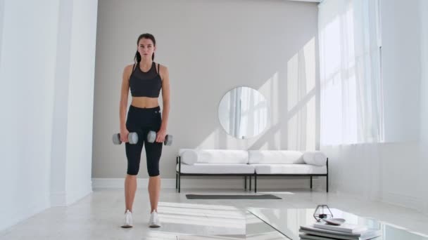 En ung atletisk kvinna i en ljus lägenhet utför en marklyft med hantlar hemma, vilket gör lutar framåt för att stärka musklerna i låren. — Stockvideo