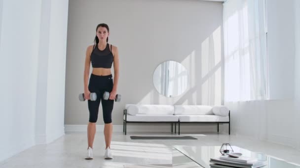 Eine junge, athletische Frau in einer hellen Wohnung führt zu Hause ein Kreuzheben mit Kurzhanteln durch, wobei sie sich nach vorne lehnt, um die Muskeln der Oberschenkel zu stärken. — Stockvideo