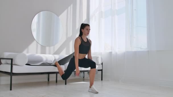 Brunette in zwarte Sportswear in een wit appartement maakt een split Squat met dumbbells in haar handen leunend op de Bank met haar voet — Stockvideo