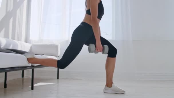 Brunette in black sportswear in a white apartment makes a split squat with dumbbells in her hands leaning on the sofa with her foot — Stock Video