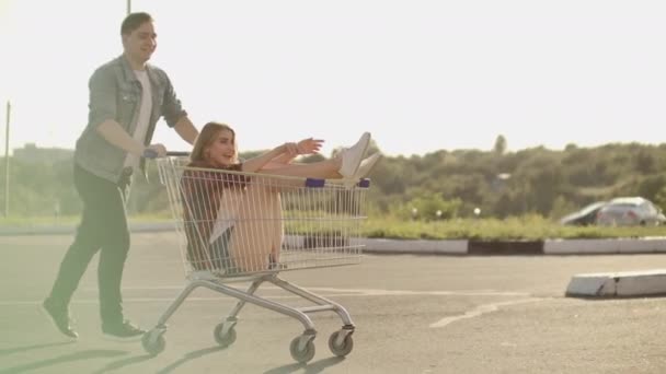 Side view of a young female and male having fun outdoors on shopping trolleys. Multiethnic young people racing on shopping carts. On the parking zone with their — Stock Video