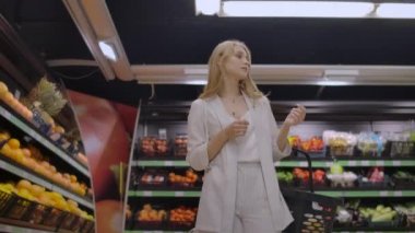 Güzel bir kadın elinde meyve raflarına bakarak bir sepet ile süpermarkette yürüyor. Taze meyve ve sebze satın alma