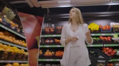 Güzel bir kadın elinde meyve raflarına bakarak bir sepet ile süpermarkette yürüyor. Taze meyve ve sebze satın alma