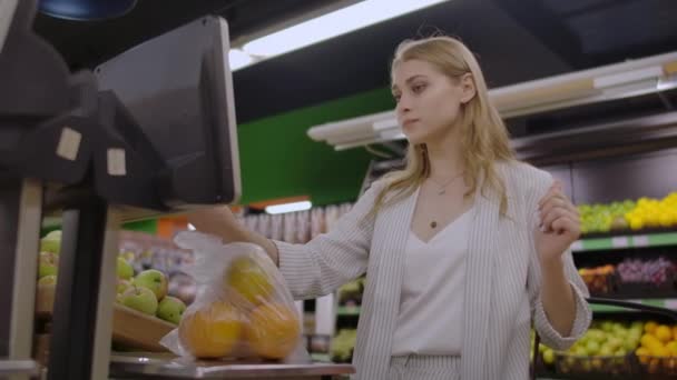 Una ragazza bionda in un supermercato pesa arance su una bilancia elettronica premendo il display in piedi con un cesto in mano . — Video Stock