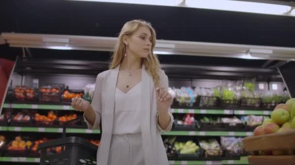 Una hermosa mujer camina por el supermercado con una cesta en las manos mirando los estantes de fruta. Compra de frutas y verduras frescas — Vídeos de Stock