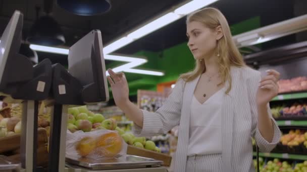 Una ragazza bionda in un supermercato pesa arance su una bilancia elettronica premendo il display in piedi con un cesto in mano . — Video Stock
