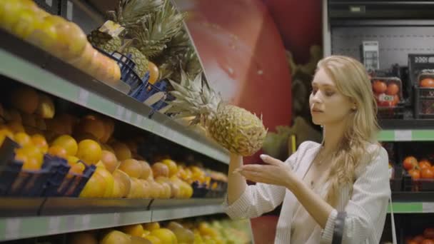 Frau wählt eine Ananasfrucht aus und legt sie in den Einkaufskorb, nimmt kleine Ananas, die in der Nähe liegt. Kundin in der Obst- und Gemüseabteilung eines großen modernen Supermarktes. — Stockvideo