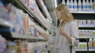 Bir kadın süpermarket buzdolabından bir şişe süt alır ve etiket ve ürünün bileşimi okur. Sepete süt koyun