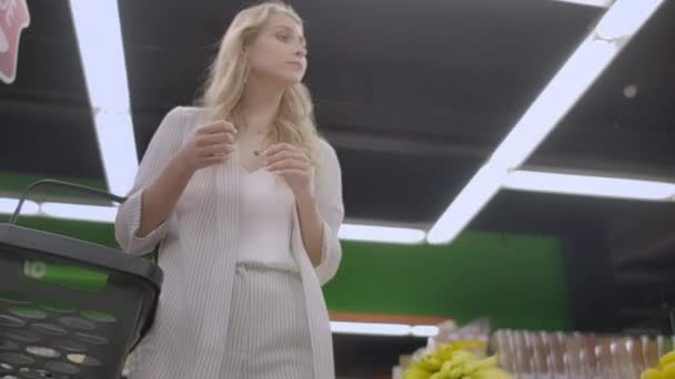 Mooie dame in casual kleding is wandelen in supermarkt sturende winkelwagentje met voedsel erin en rondkijken op de planken met producten. Vrouwen en winkels concept — Stockvideo