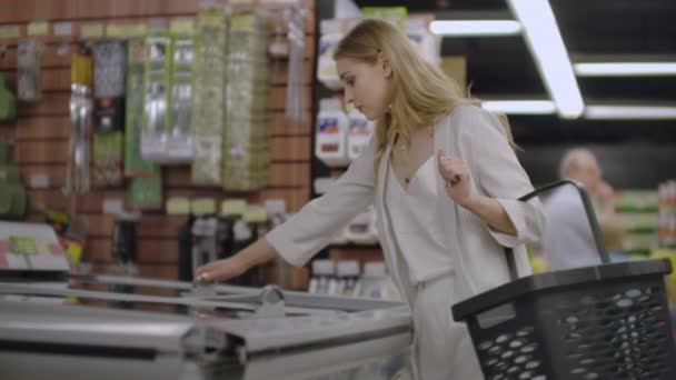 A menina no supermercado tira da geladeira alimentos congelados lê a composição do produto e coloca-o na cesta . — Vídeo de Stock