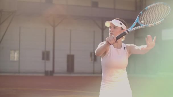 En professionell kvinna i en vit tight kostym träffar bollen med ett racket och dynamiskt spelar på tennisbanan — Stockvideo