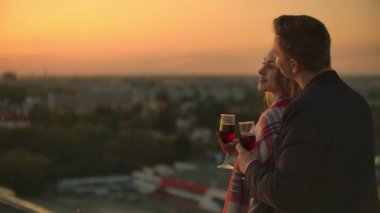 Erkek ve genç güzel kadın kanepede oturan, şarap bardakları tutan ve gün batımında çatı terasında öpüşme.