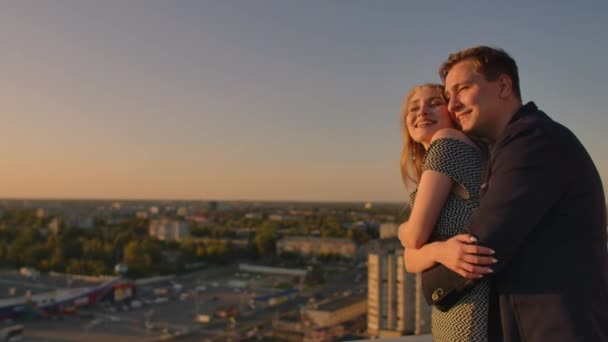 Encantador casal feliz doce sentar-se no topo do telhado com incrível vista para o pôr do sol na cidade urbana da cidade. Eles amam uns aos outros abraços muito ternura sol vai entre o cabelo . — Vídeo de Stock